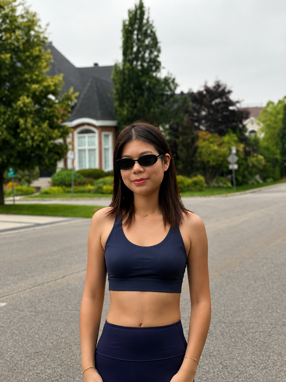 A woman wearing a navy blue sports bra and matching high-waisted leggings stands on an asphalt street in front of a suburban house with trees.